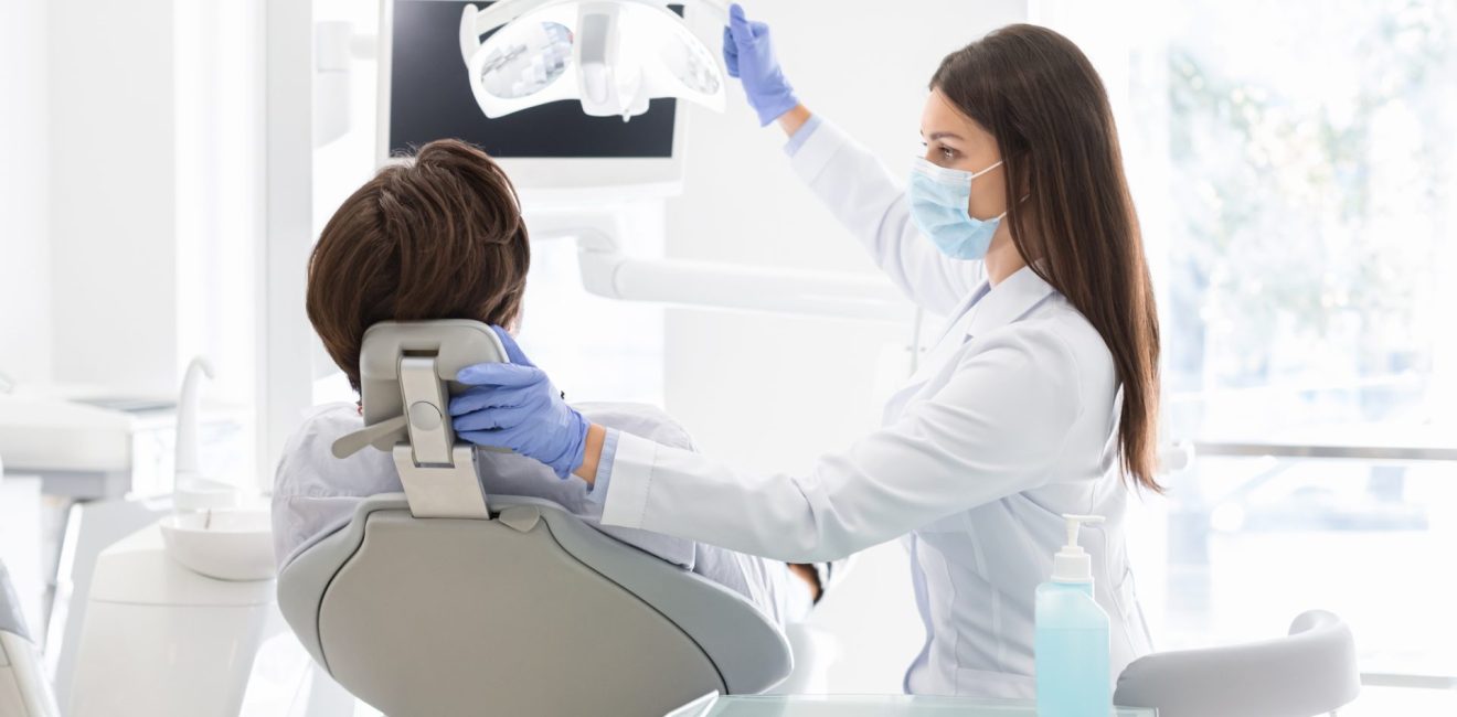 Female dentist doctor greeting patient sitting in dentist chair, turning lamp on, copy space