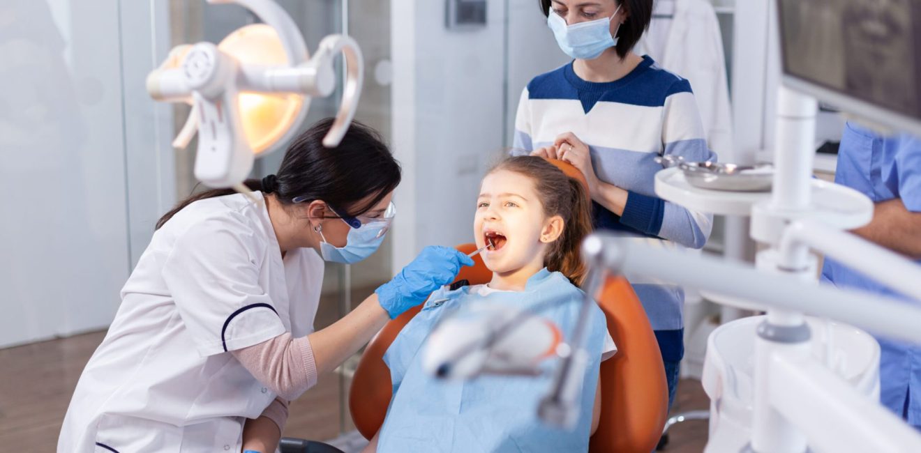 Angled mirror used by dentist doctor on little girl with mouth open in dental office. Dentistry specialist during child cavity consultation in stomatology office using modern technology.