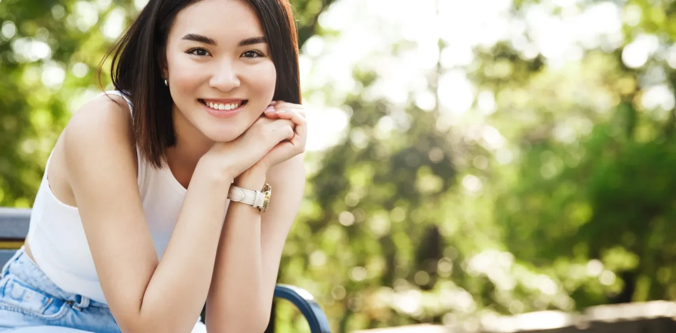 Beautiful asian woman sitting on bench and smiling. Modern girl resting in park on sunny summer day, looking happy.